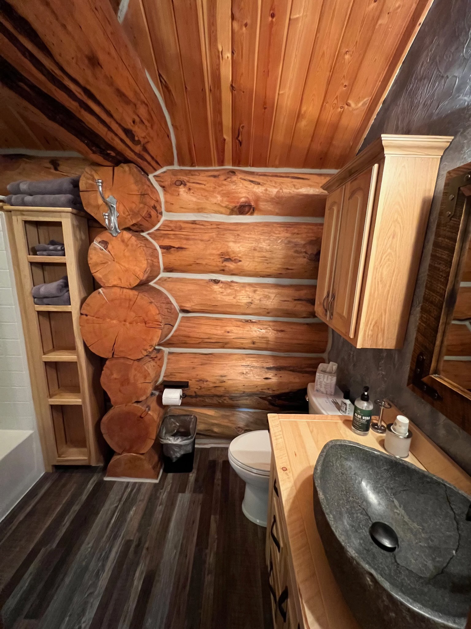 Rustic ensuite bath with a river-stone vessel sink, log cabin walls, and a wood plank ceiling
