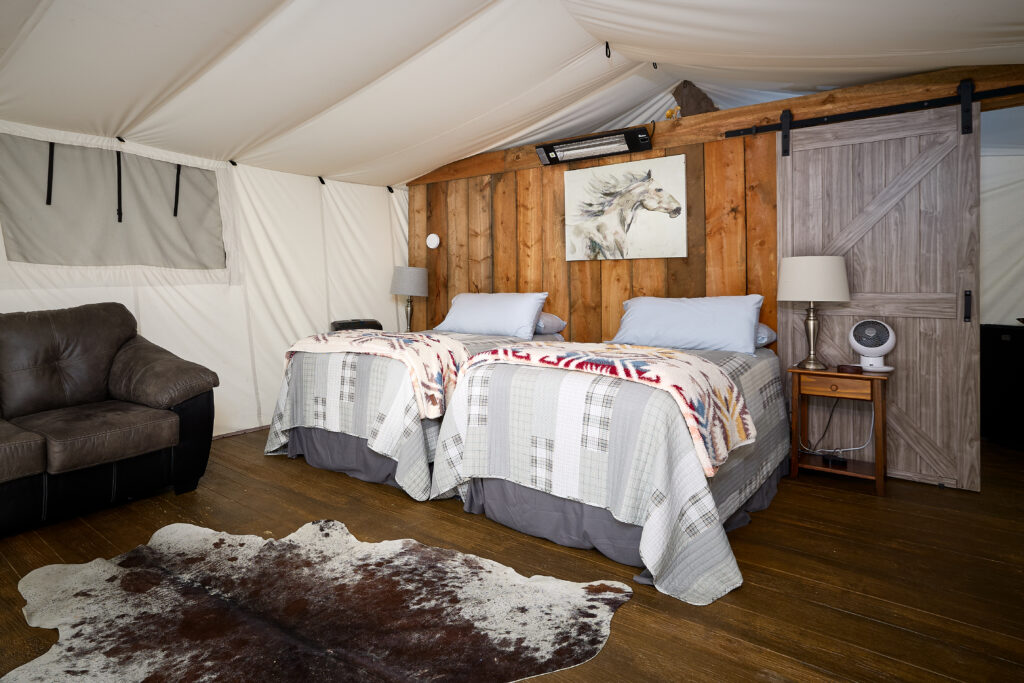 Two twin beds with plaid quilts and Pendleton-style throws sit against a wood-paneled wall inside a canvas tent room.