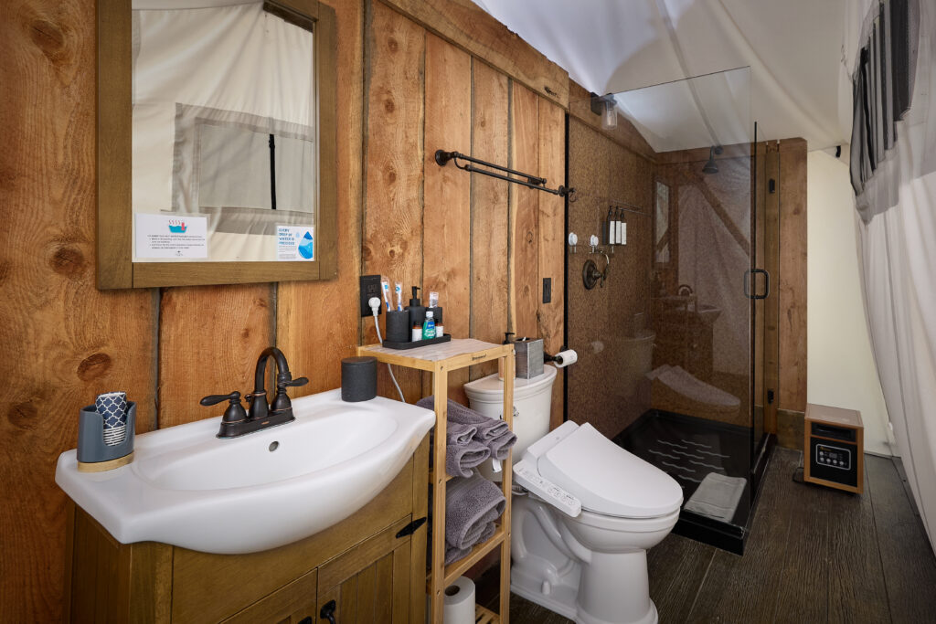 Wood vanity sink with bronze fixtures and a wood-framed mirror beside a glass shower and bidet toilet in a tent bathroom