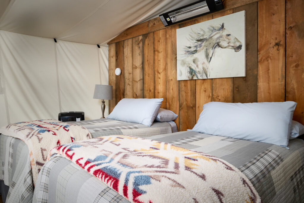 Crisp white pillows and folded Pendleton-style blankets top two twin beds beneath a horse painting on wood paneling.
