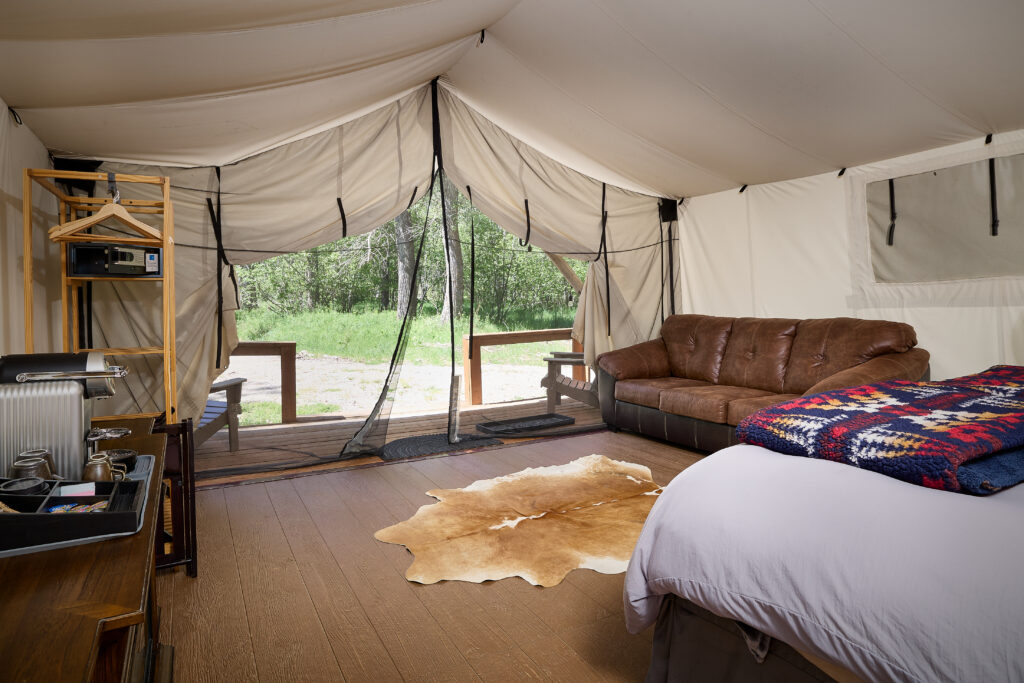Mesh screen doors open from a canvas tent lounge area onto a wooded deck, with a leather sofa and cowhide rug inside.