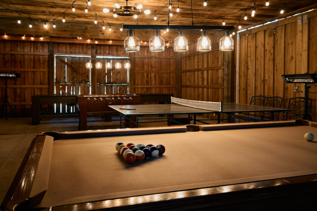 A pool table with balls racked sits in a rustic wood-paneled game barn, string lights overhead, ping pong table behind