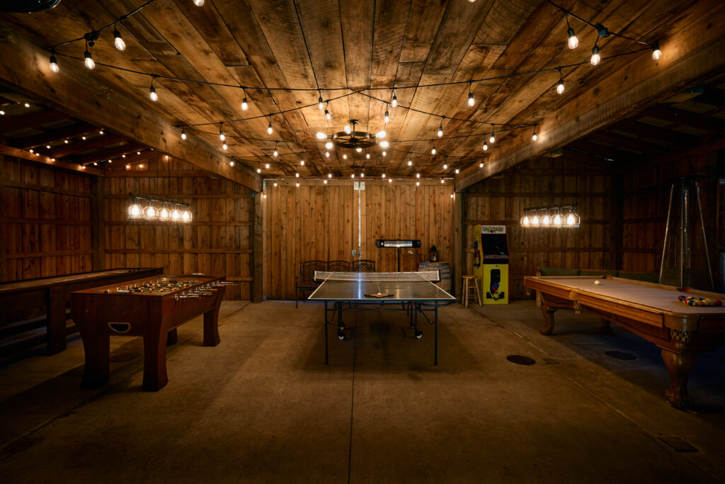 Foosball, ping pong, and pool tables under string lights, with an arcade cabinet glowing in the wood-plank rec barn