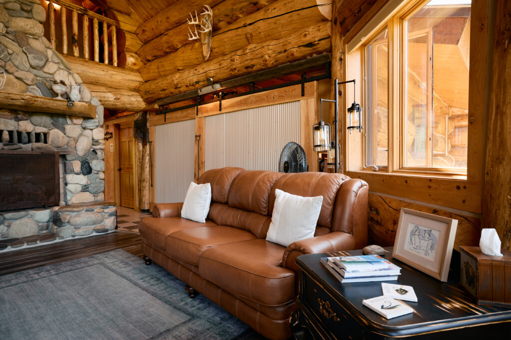 A leather sofa with white throw pillows sits beside the stone fireplace in a cozy log-cabin corner with wall lanterns.
