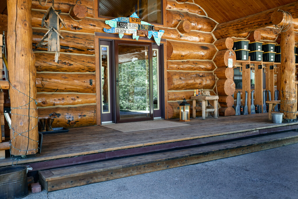 Covered log lodge entry porch with fish-shaped license-plate art above the door and waders hanging along the rail.