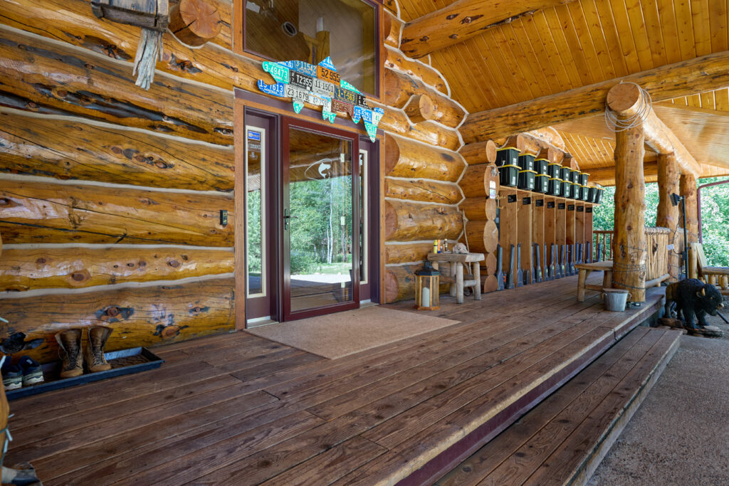 A long covered porch lined with hanging waders and boots beneath the lodge's log walls, leading to the glass entry door.