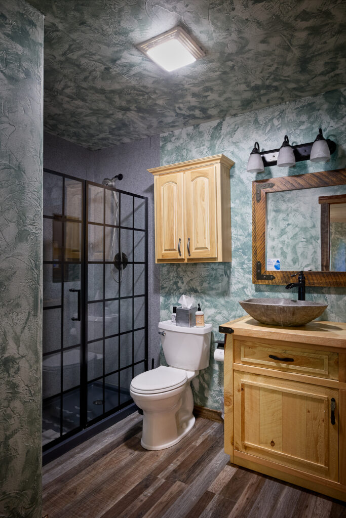 An ensuite bathroom with a glass-paneled shower, a stone vessel sink, and a rustic wood-framed mirror.