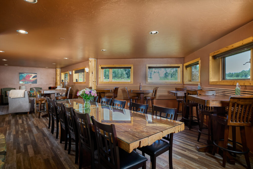 A long dining room with a rustic wood table set for a large group, flanked by windows overlooking greenery.