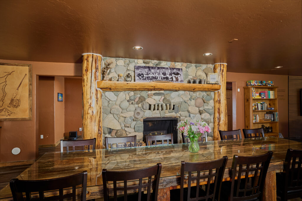 A great room with a large river-stone fireplace and mantel, seen behind a long dining table set with fresh flowers.