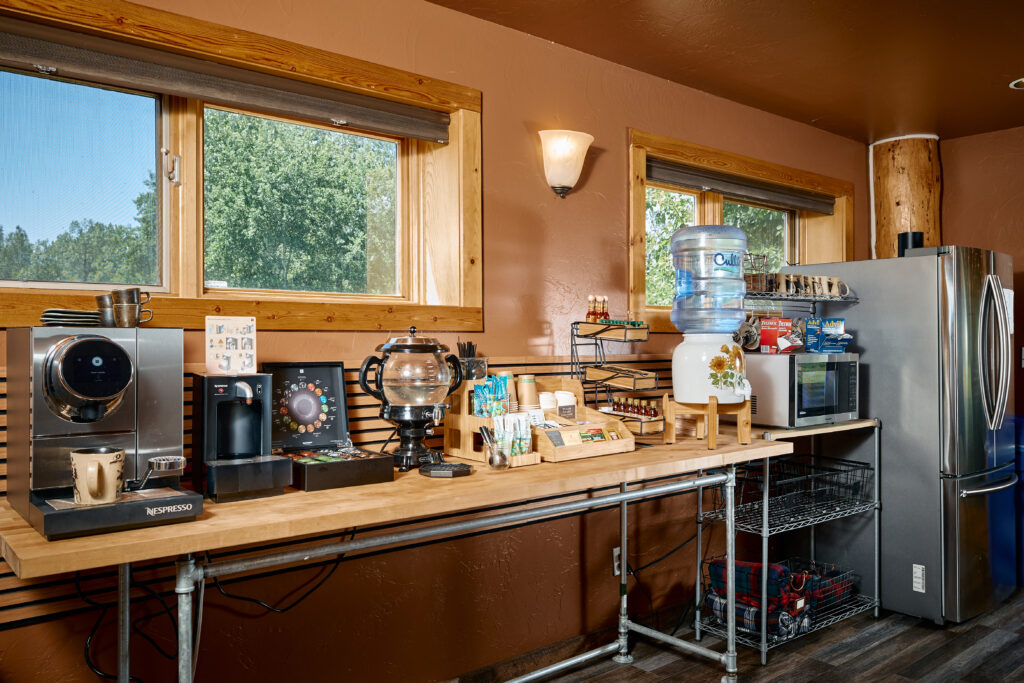 A coffee station with an espresso machine, bottled water cooler, mini fridge, and snacks on a wood counter.