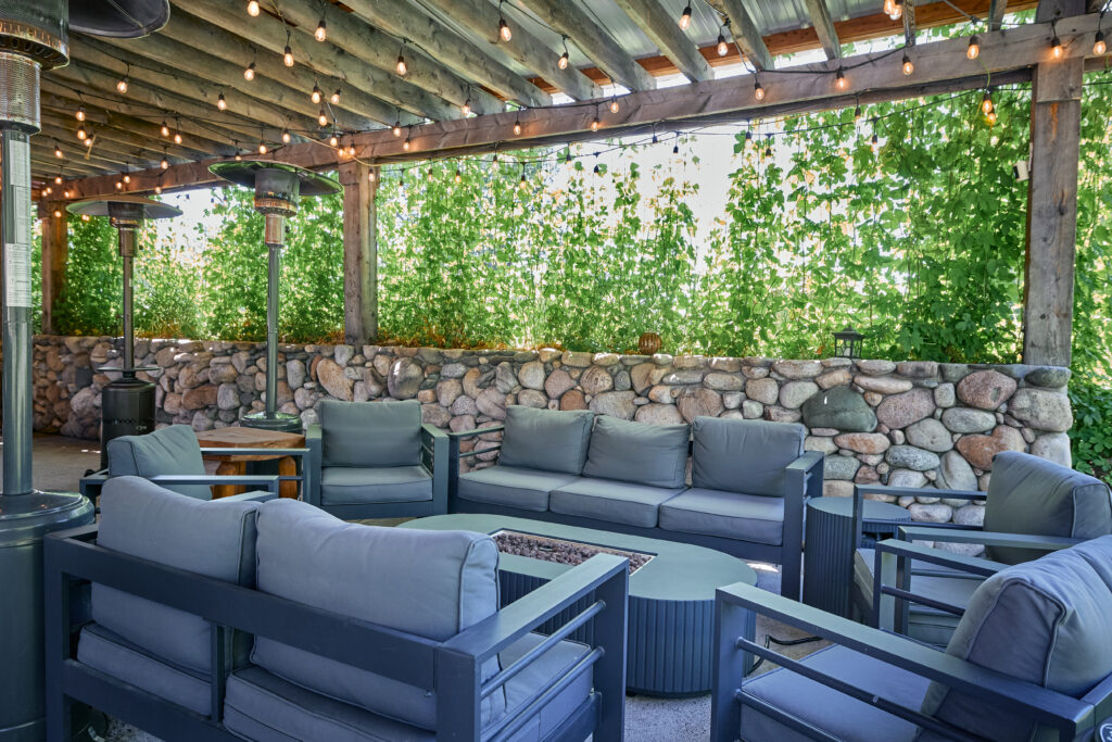 An outdoor lounge with blue-gray sofas around a built-in fire pit table, beneath a string-lit pergola and hop vines.