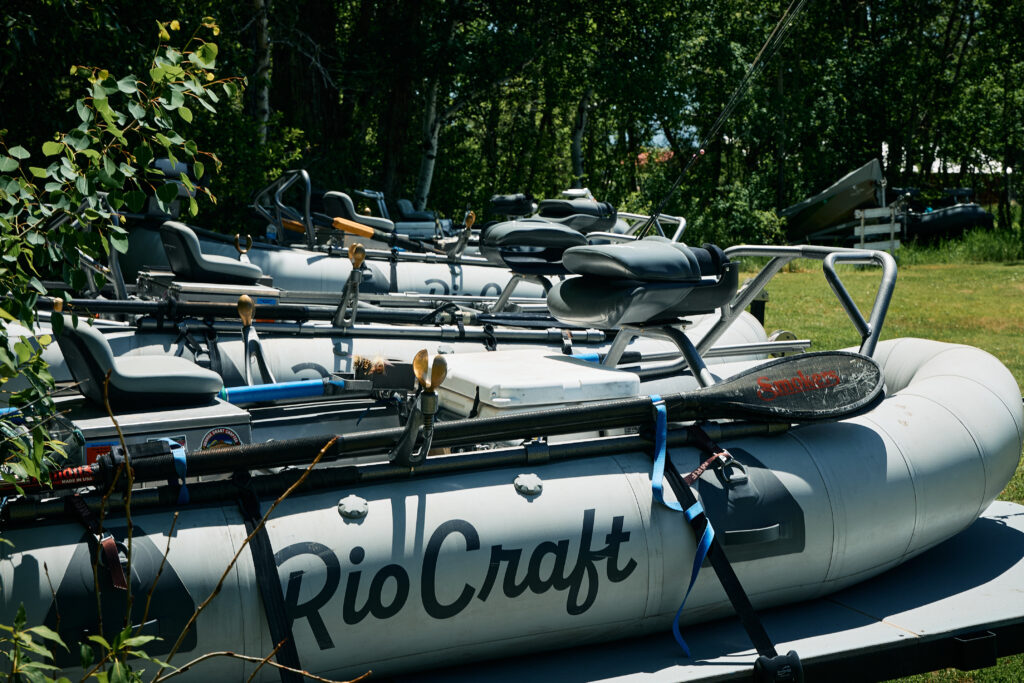 Fleet of white RioCraft drift boats rigged with rod holders, oars, and a cooler, parked in the shade near the lodge