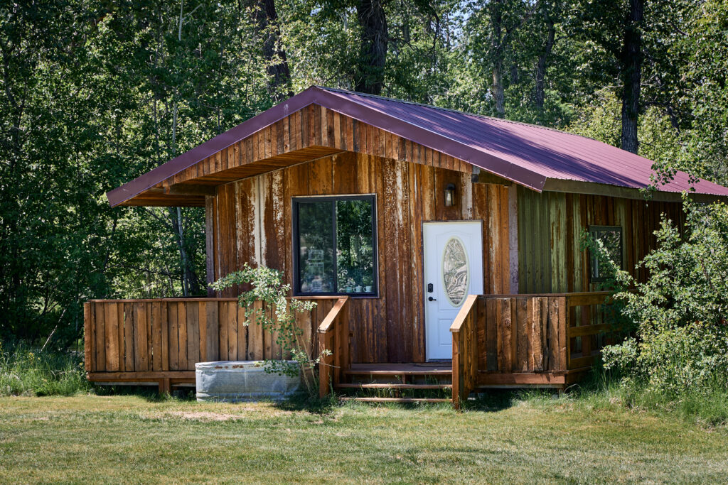 A small guest cabin with weathered wood siding and a dark red metal roof, tucked among trees on a grassy lawn