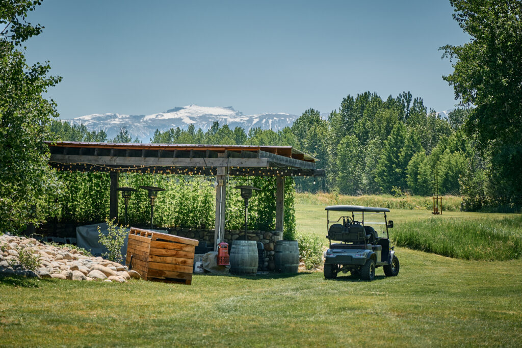 A covered outdoor bar on a stacked-stone and barrel base, with patio heaters, golf cart parked nearby, mountains beyond
