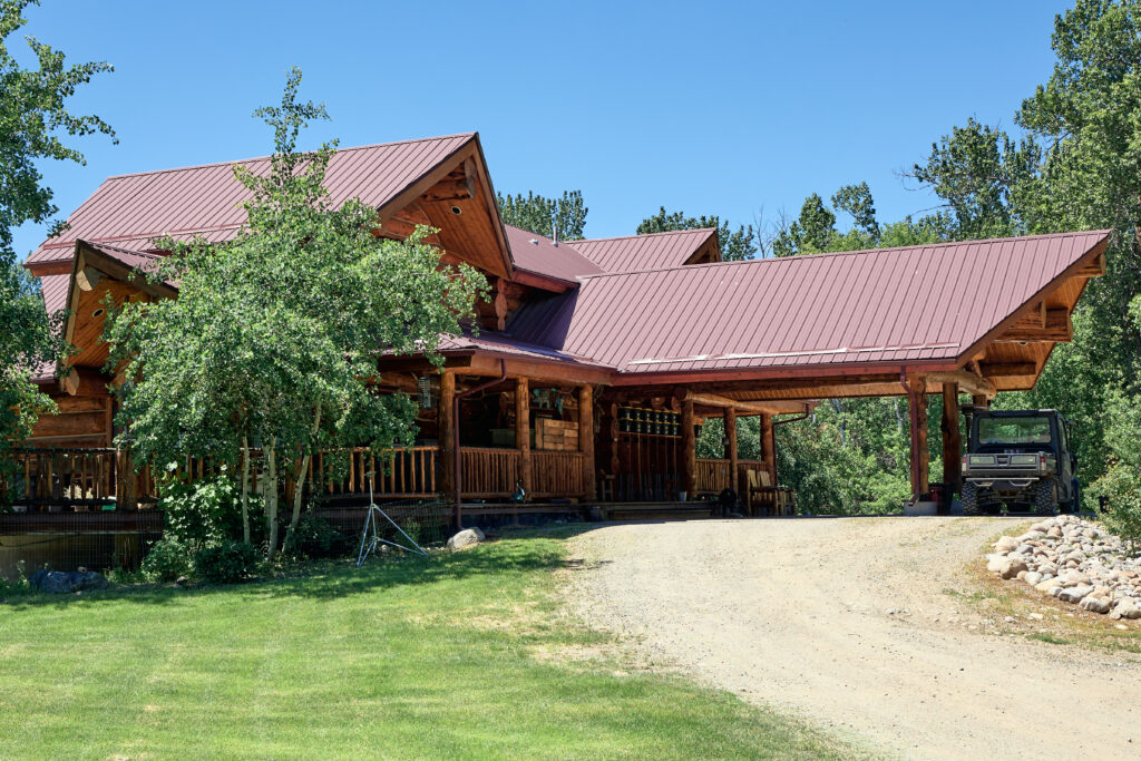 A UTV parked beneath the lodge's log-post breezeway, dark maroon metal roofline over the curving gravel driveway