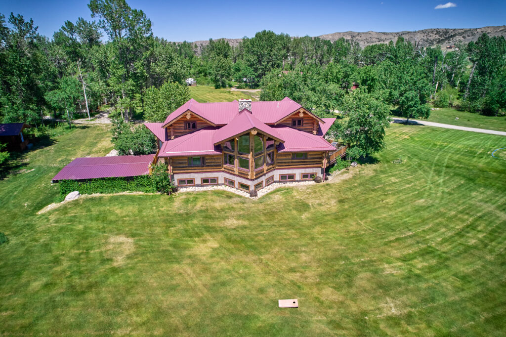 The log lodge's maroon roofline and glass great-room gable above a mowed lawn ringed by trees and distant bluffs