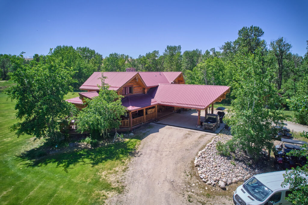 Gravel drive curves past rock landscaping to the lodge's carport, where a UTV waits beside golf carts parked under trees