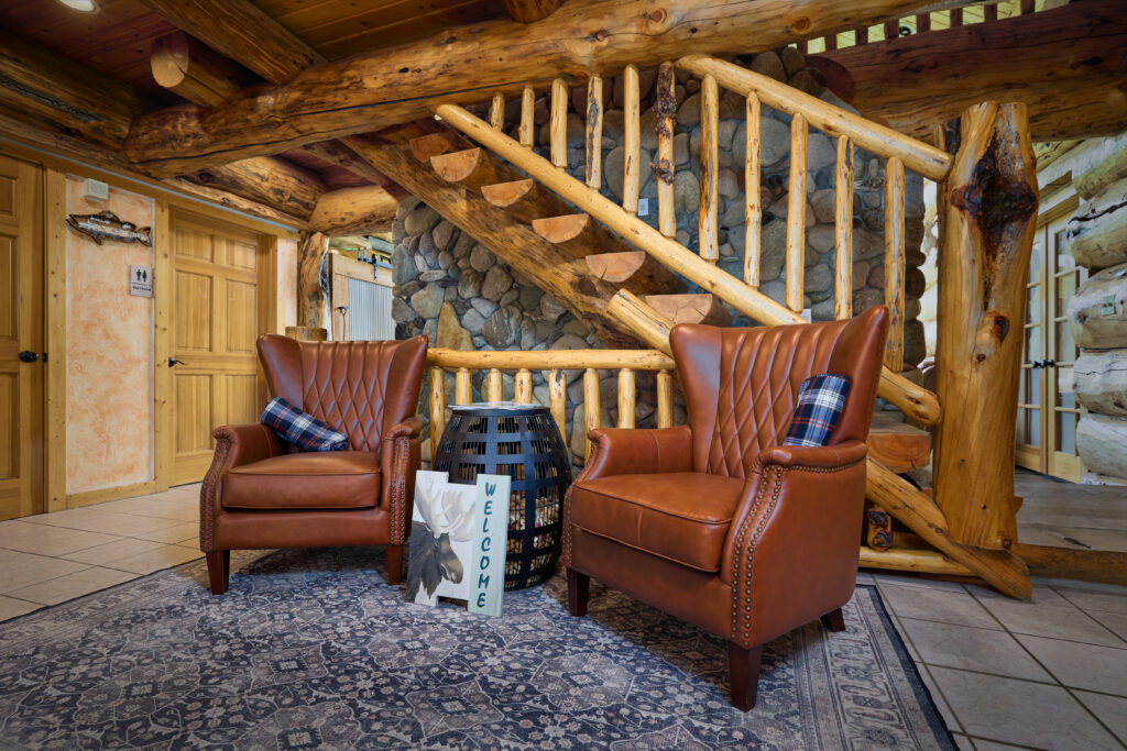 Two leather armchairs and a moose-themed welcome sign sit beneath the lodge's log staircase