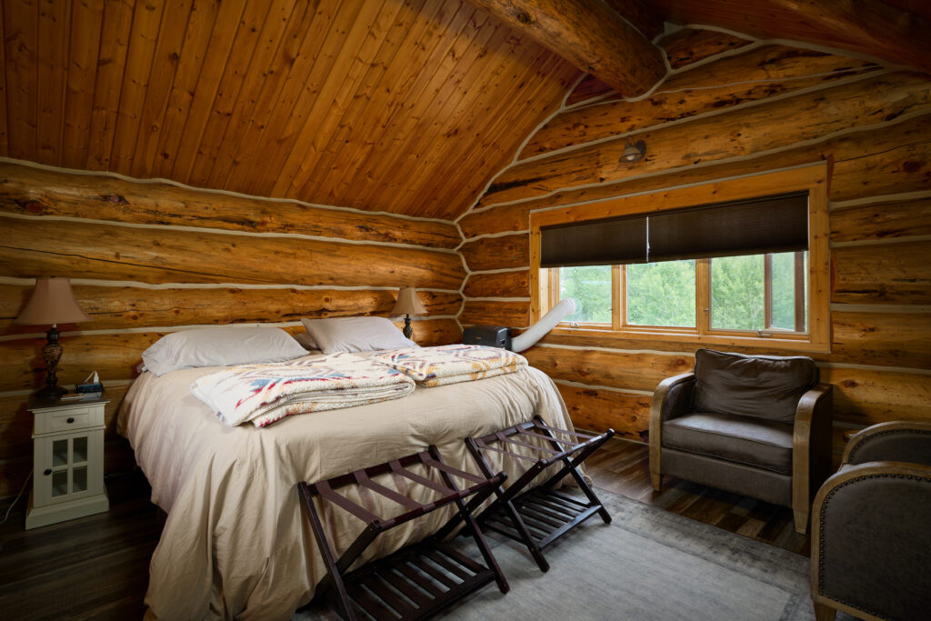 A log-walled bedroom with vaulted wood ceiling, a made bed, and two upholstered armchairs by the window