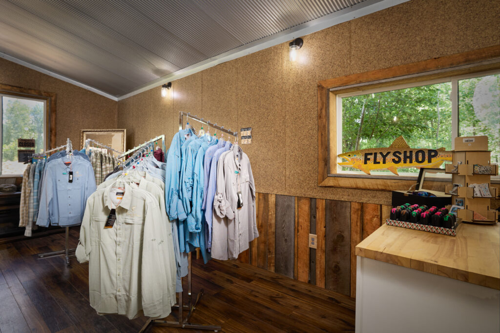 A wooden fish-shaped Fly Shop sign hangs above racks of shirts in the lodge's retail shop