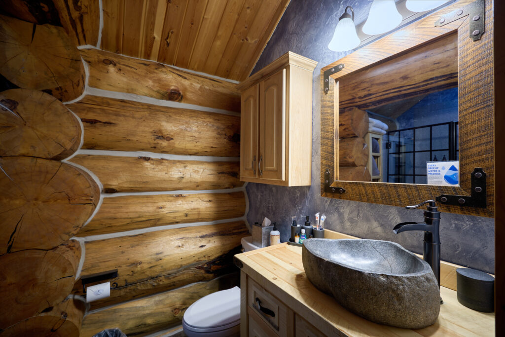 River-stone vessel sink and matte-black faucet beside a log wall, below a log-slab mirror with riveted metal strapping
