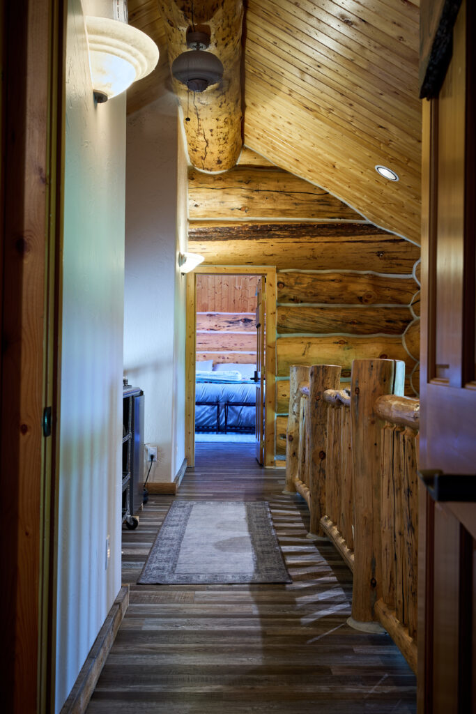 An upstairs loft hallway with a log-pole railing leading past pendant lights toward a bedroom