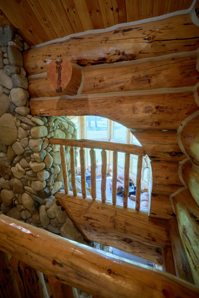 A view through log timber beams and a river-stone column down to a great room with seating below