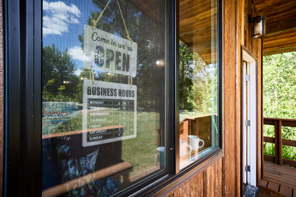 A screened porch window displaying an "Open" sign and posted business hours amid summer greenery