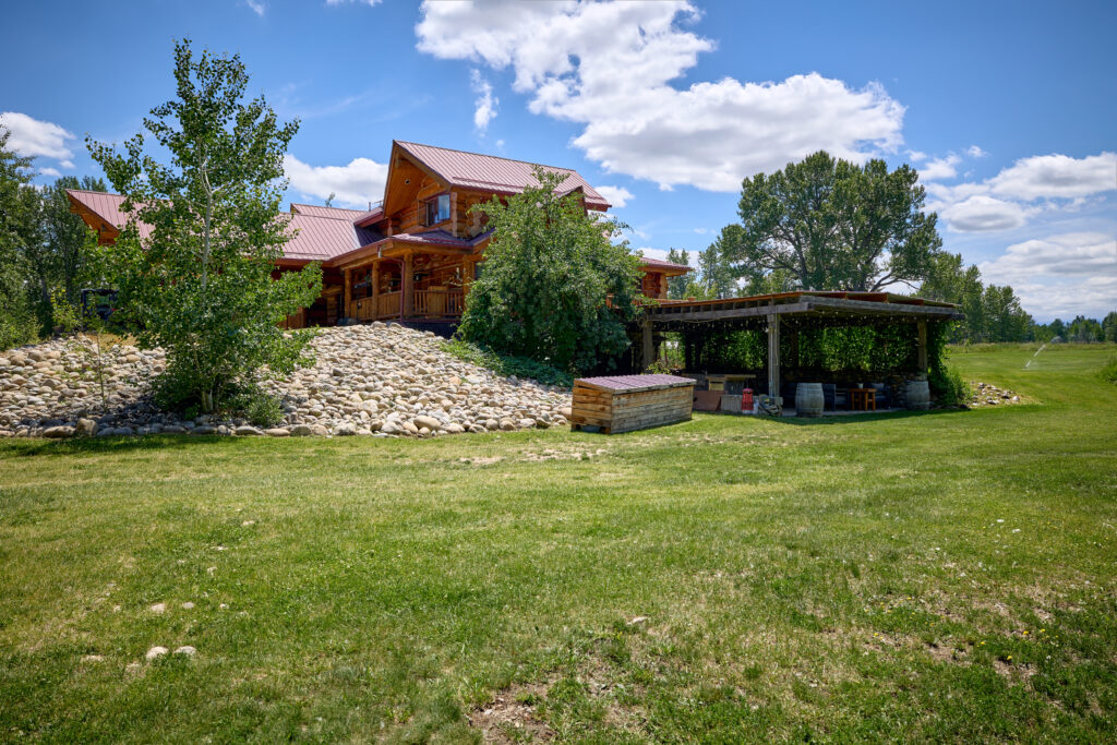 Pergola patio with wine barrels and string lights beside a river-rock landscaped mound at the log lodge