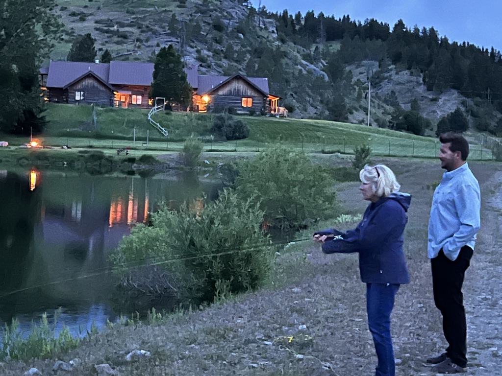 Two anglers casting on the trout pond at dusk with the lit lodge reflected in the water